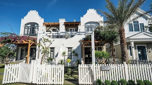 A white building with a white picket fence, palm trees, and a pergola with purple flowers.