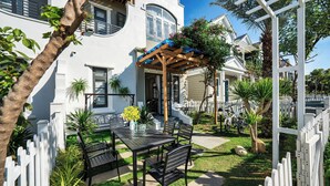A patio with a table and chairs, surrounded by a white picket fence and various plants.