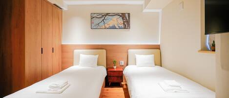 A hotel room with two single beds, wooden wardrobe, a painting on the wall, and a TV mounted on the wall.