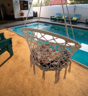 Ein Hängestuhl aus Makramee mit Kissen, ein Pool und im Hintergrund grüne Liegestühle.