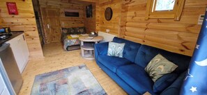 Interior de uma cabana de madeira com um sofá, uma mesa pequena, uma cama com colcha florida e uma área de cozinha.