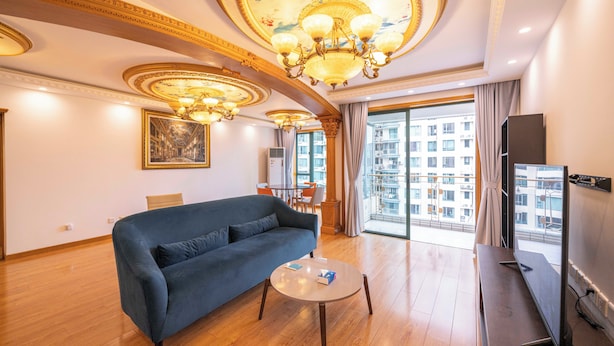 A spacious living room with a blue sofa, a round coffee table, and a flat-screen TV. The room features ornate ceiling decorations and a large window with a city view. - Tyms Xujiahui Premium 2BR APT Metro (Shanghai)