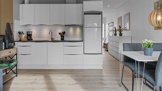 A modern kitchen with white cabinets, a built-in oven, and a coffee machine. There is a dining area with a table and chairs, and a plant on the table.
