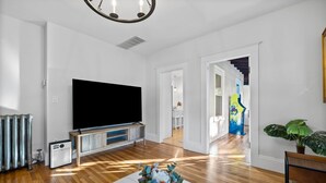 Amplia sala de estar con una gran televisión de pantalla plana sobre un soporte de madera, un radiador y una planta en maceta.