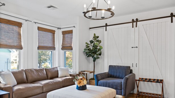 Sala de estar con sofá de cuero marrón, un sillón azul, un otoman blanco y un banco de madera. Hay una planta grande en una maceta azul, un candelabro y una puerta corrediza de estilo rústico.