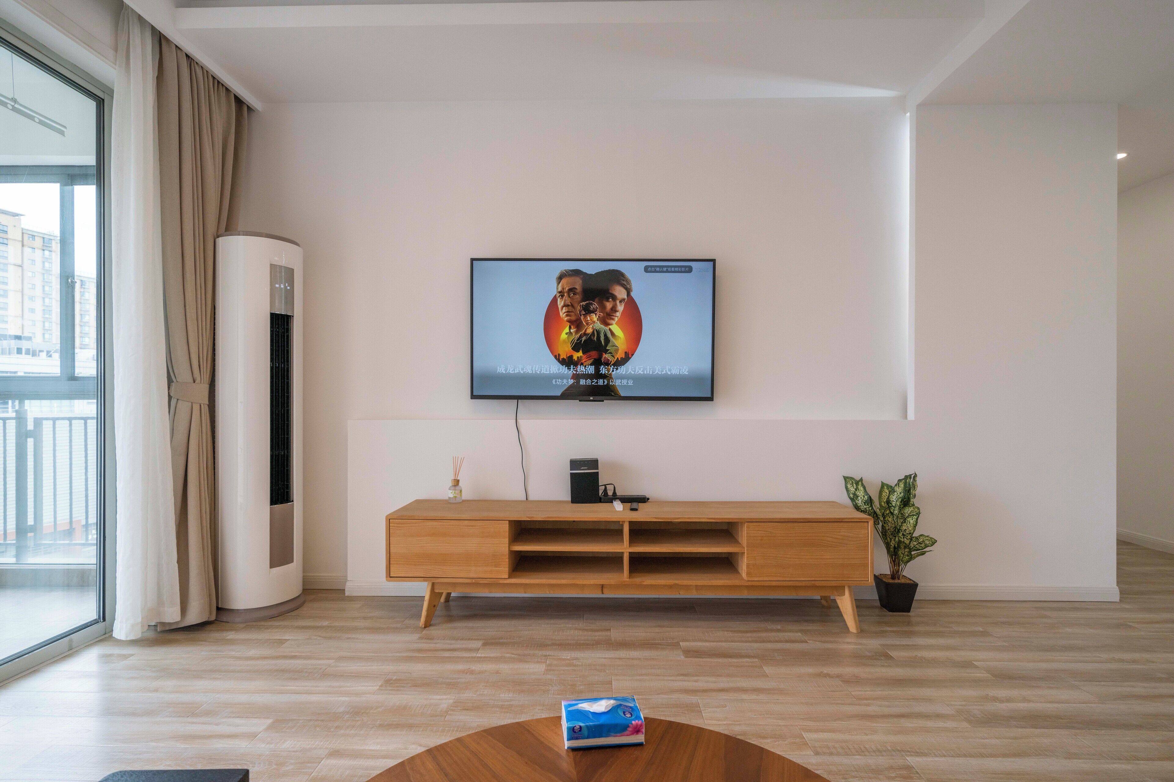 A modern living room with a flat-screen TV mounted on the wall, a wooden entertainment unit below, and a potted plant to the right.