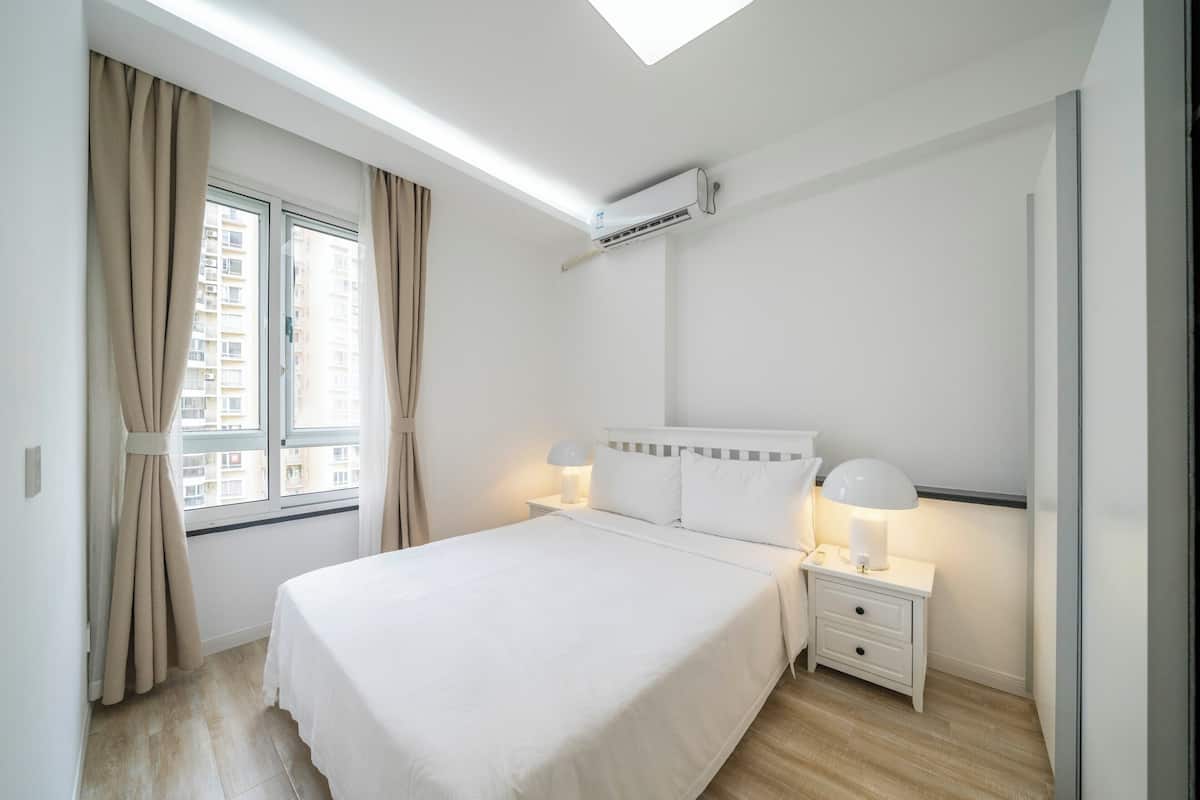 A modern bedroom with a large bed, bedside lamps, a window with curtains, and an air conditioning unit.