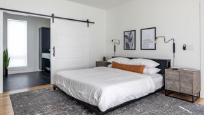 A modern bedroom with a large bed, a wooden nightstand, a wall-mounted lamp, and a framed quote on the wall. - Modern Oasis: Rooftop Sunsets & City Lights! (Nashville)
