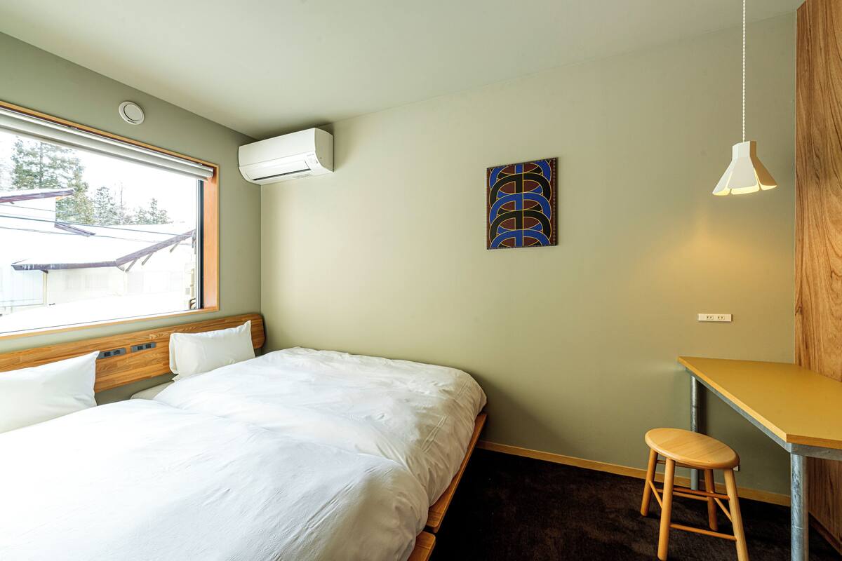Un dormitorio con cama, escritorio, taburete, aire acondicionado, un cuadro y ventana.