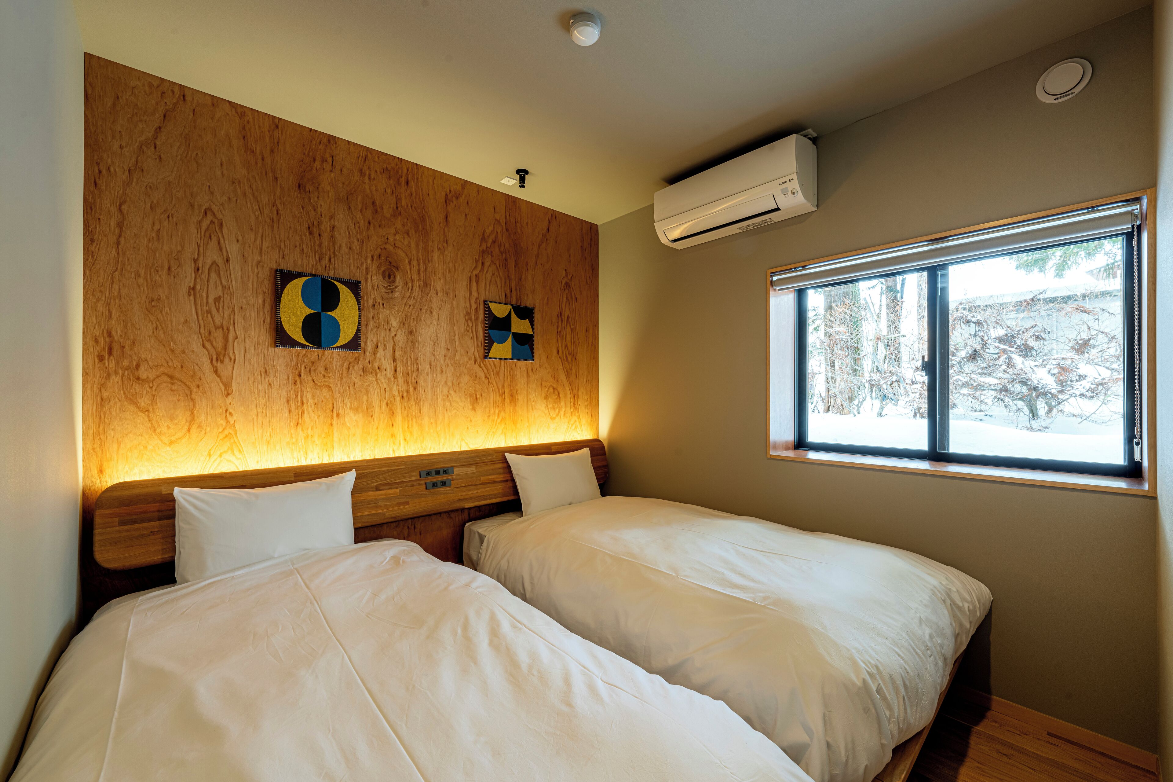 A room with two beds, wooden headboards, a large window with a view of trees, and an air conditioning unit.