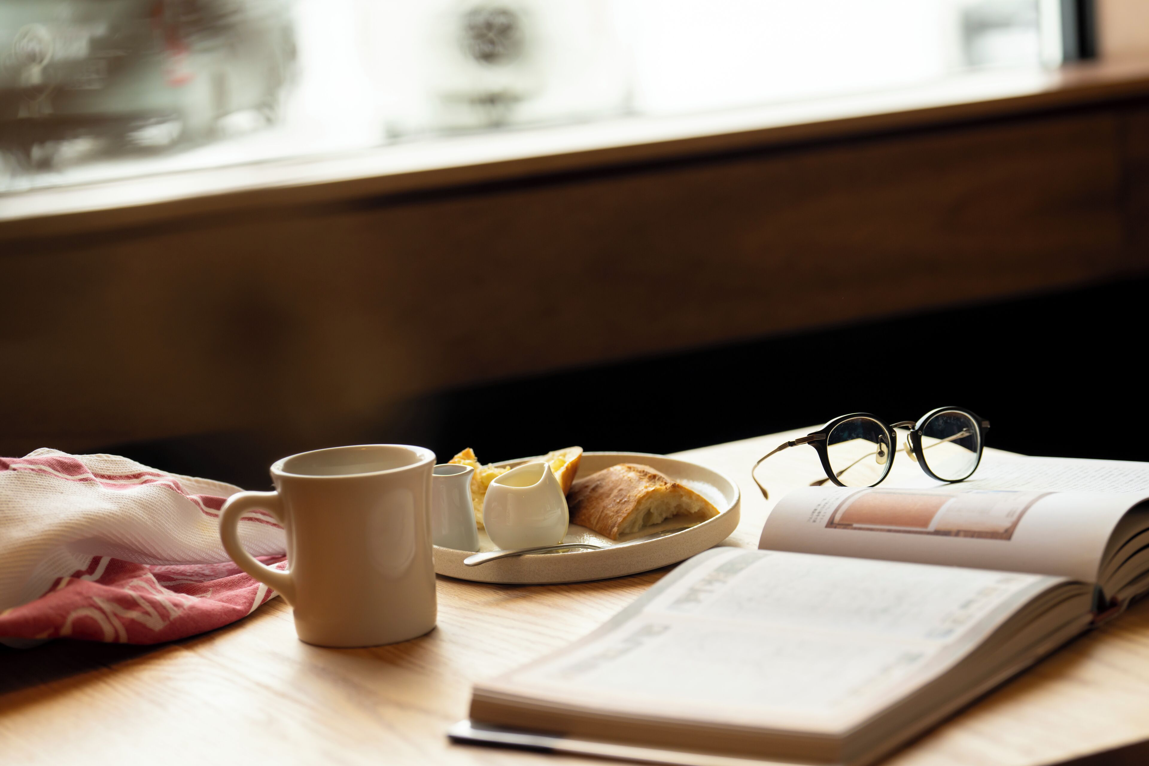 Una mesa con una taza, un plato de pan y un recipiente pequeño, un par de gafas y un libro abierto.