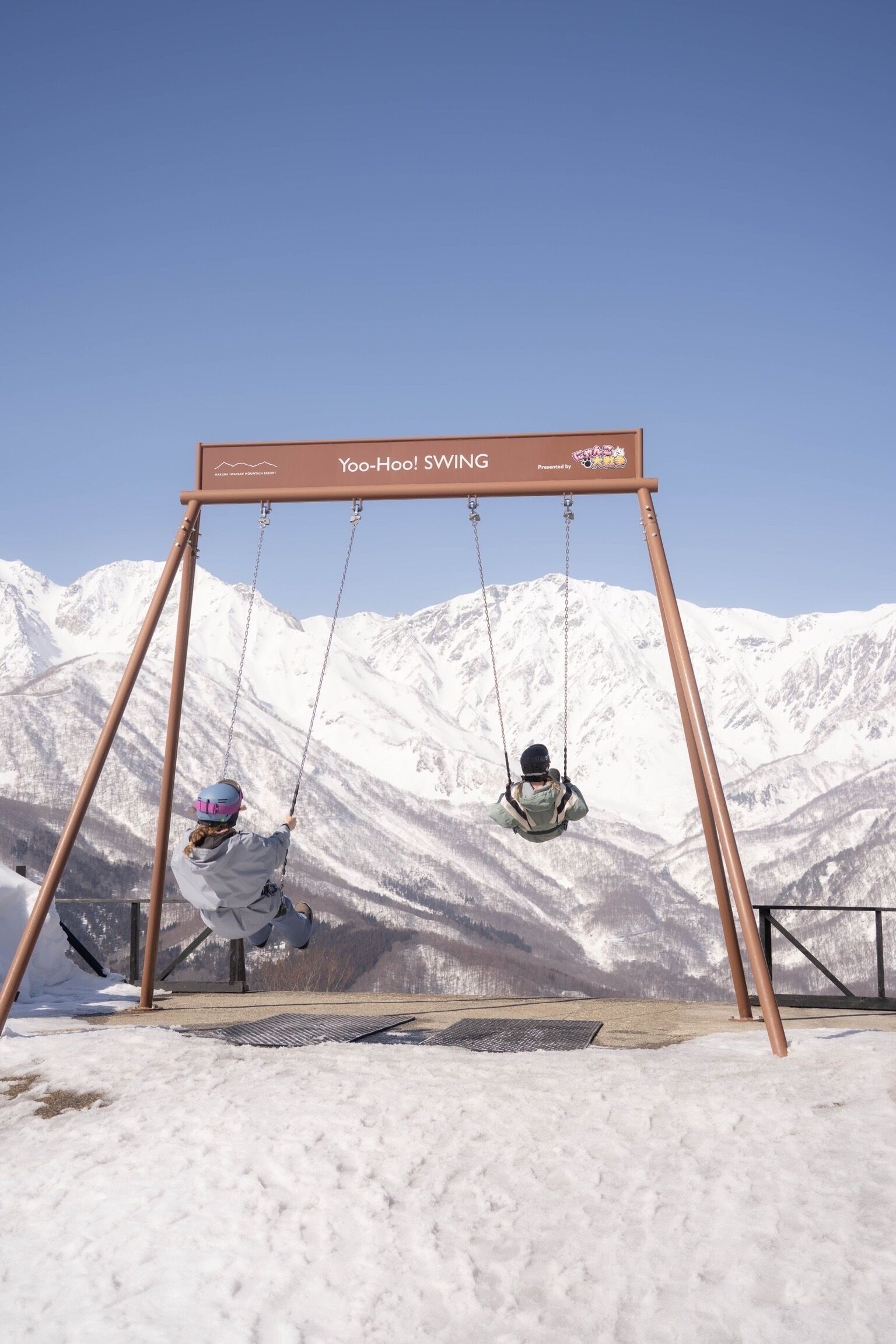 A swing set with a sign reading 'Yoo-Hoo! SWINGING' in a snowy mountainous area.