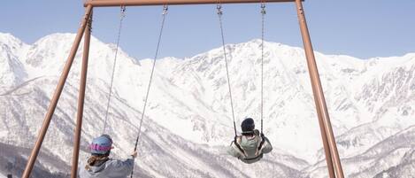 A swing set with a sign reading 'Yoo-Hoo! SWINGING' in a snowy mountainous area.