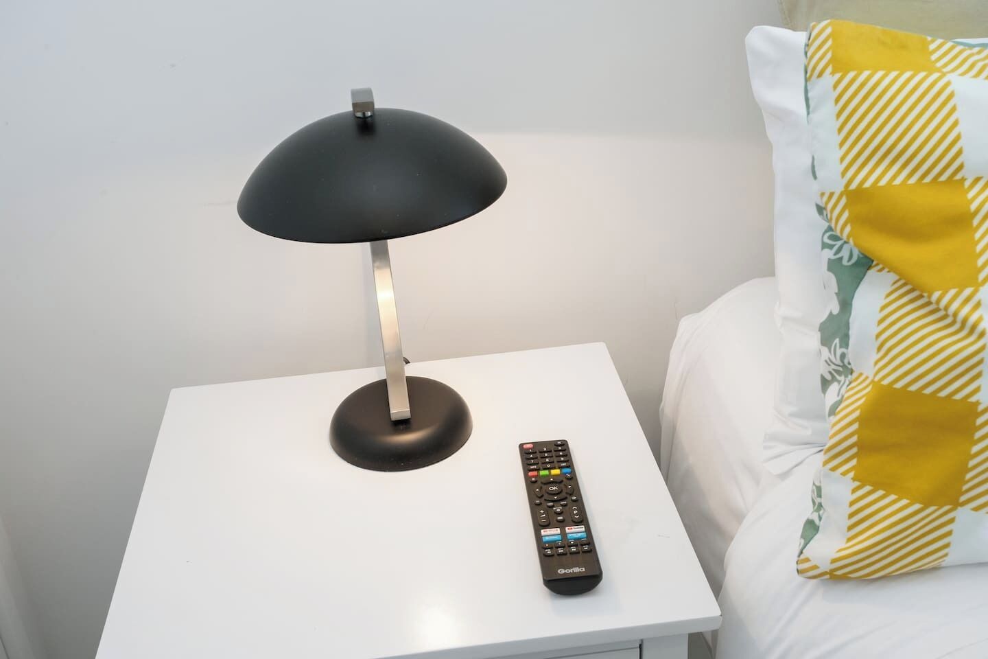 Una mesita de noche blanca con una lámpara negra y un control remoto.
