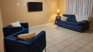 Una sala de estar con dos sofás azules, un televisor de pantalla plana, una lámpara y un ventanal con cortinas.