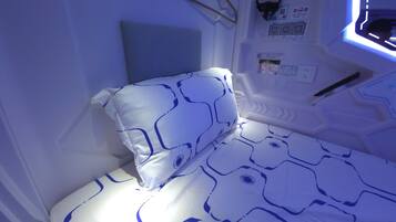 A small, single-bed room with a blue and white patterned bedspread, a pillow, and a headboard. There are wall-mounted controls and a window with a view of the outside.