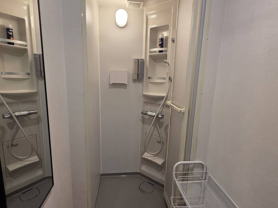 A narrow bathroom with a shower, toilet, and sink. There are shelves with toiletries and a small trash bin.