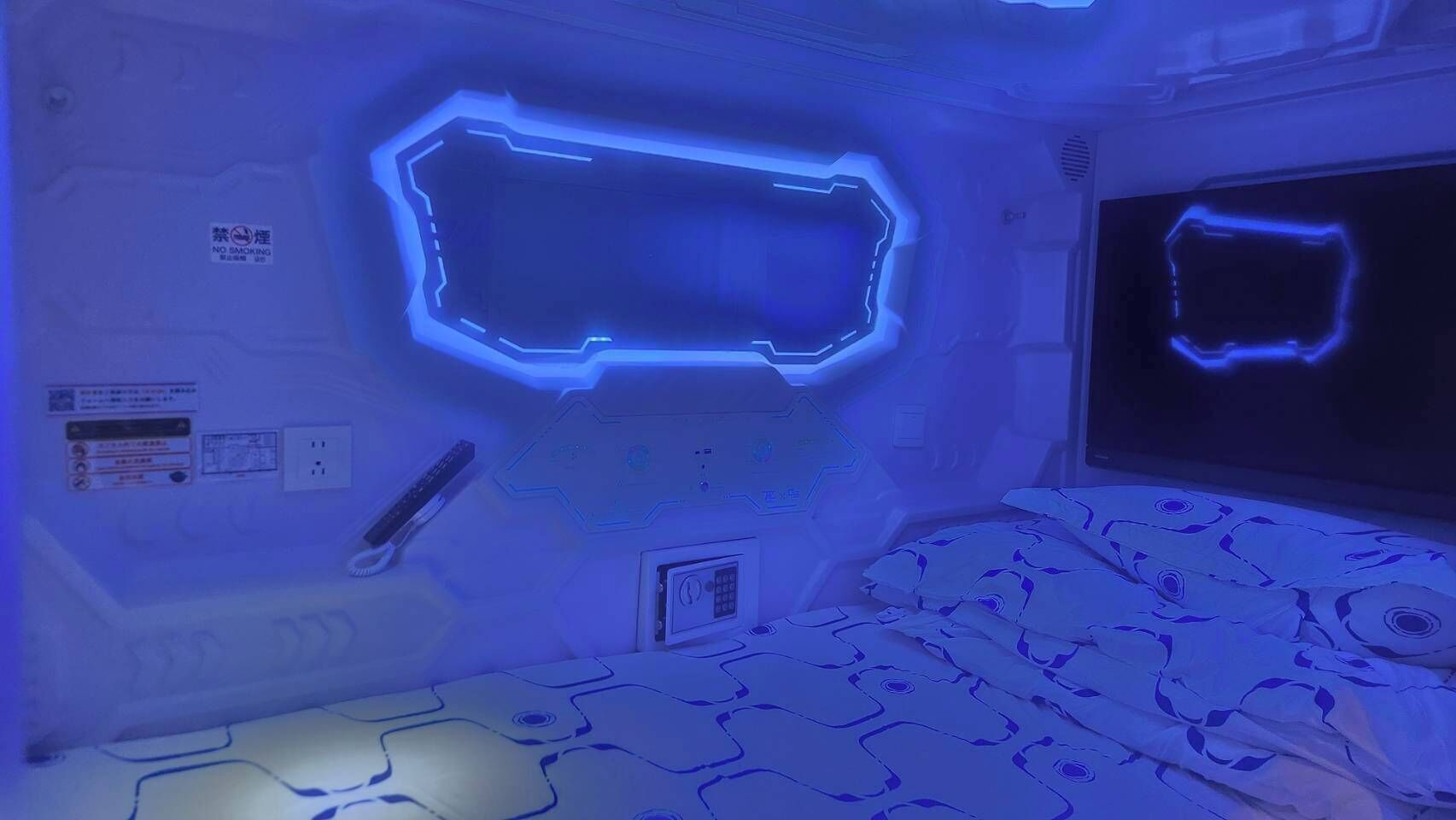 A room with a futuristic design, featuring a neon-lit geometric wall panel and a bed with a patterned cover.