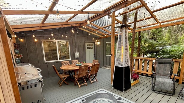 A covered outdoor deck with a hot tub, wooden furniture, and string lights. - Charming 2-bedroom cottage with a hot tub. 5 Min to Volcanoes National Park! (Volcano)