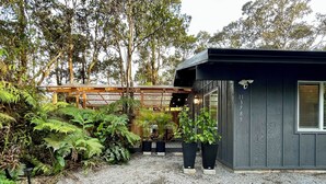 A small, dark-colored building with a gravel driveway and lush greenery. - Charming 2-bedroom cottage with a hot tub. 5 Min to Volcanoes National Park! (Volcano)