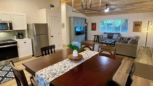 A modern kitchen with white cabinets, a stainless steel refrigerator, and a microwave. There is a dining area with a wooden table and chairs, and a living room with a gray sofa and a coffee table. - Charming 2-bedroom cottage with a hot tub. 5 Min to Volcanoes National Park! (Volcano)