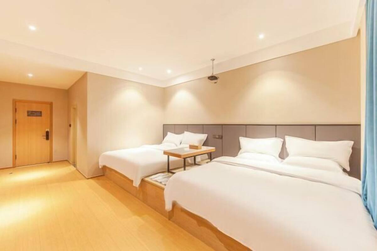 A hotel room with two beds, a wooden floor, a ceiling light, and a door.