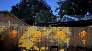 A wooden fence with four light fixtures, two chairs, and a small table in a backyard. - The White Door Studio. (Lutz)