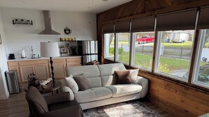 A cozy living room with a sofa, armchair, and a coffee table. A kitchen is visible in the background with a refrigerator, stove, and cabinets. Large windows offer a view of the outside. - The White Door Studio. (Lutz)