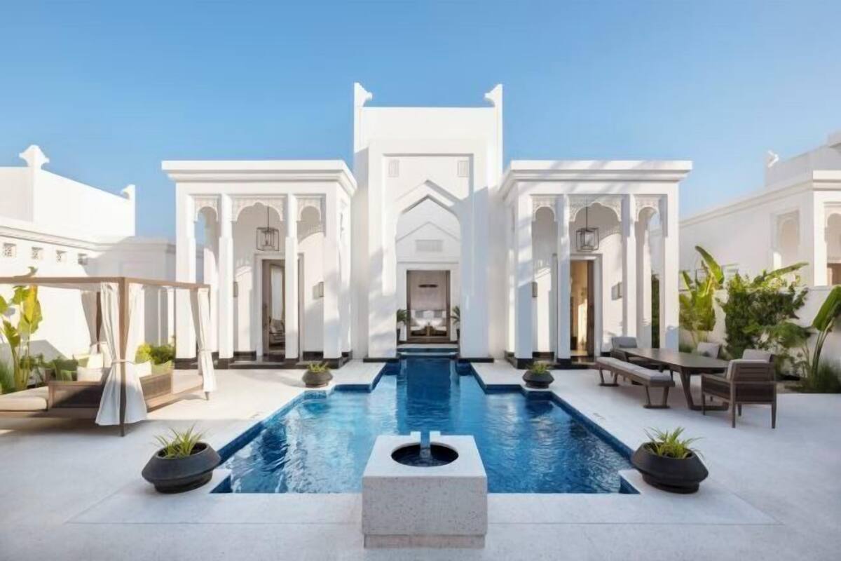 A pool area with a central water feature, surrounded by white columns and arches, with outdoor seating and plants.
