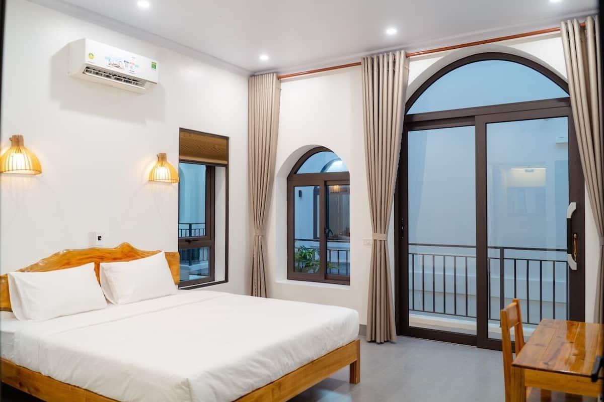 A hotel room with a bed, a wooden table, and a balcony with curtains.