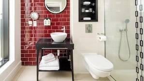 Een moderne badkamer met een rode betegelde muur, een witte wastafel, een ronde spiegel, een toilet en een douchecabine met glazen wanden.