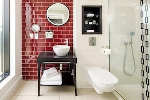 A modern bathroom with a red-tiled wall, a white sink, a round mirror, a toilet, and a glass-enclosed shower. - Hotel Indigo NEWCASTLE by IHG (Newcastle)