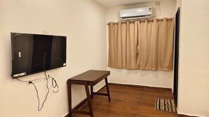 Ein Zimmer mit Flachbildfernseher an der Wand, einem Holztisch, beige farbenen Vorhängen, einer Klimaanlage und einem kleinen Teppich.