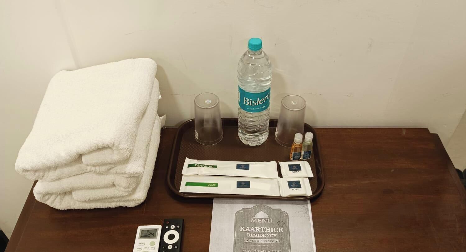 Habitación de hotel con una pila de toallas blancas, una botella de agua Bisleri, dos vasos, una bandeja con artículos de higiene bucal, un control remoto y un menú.