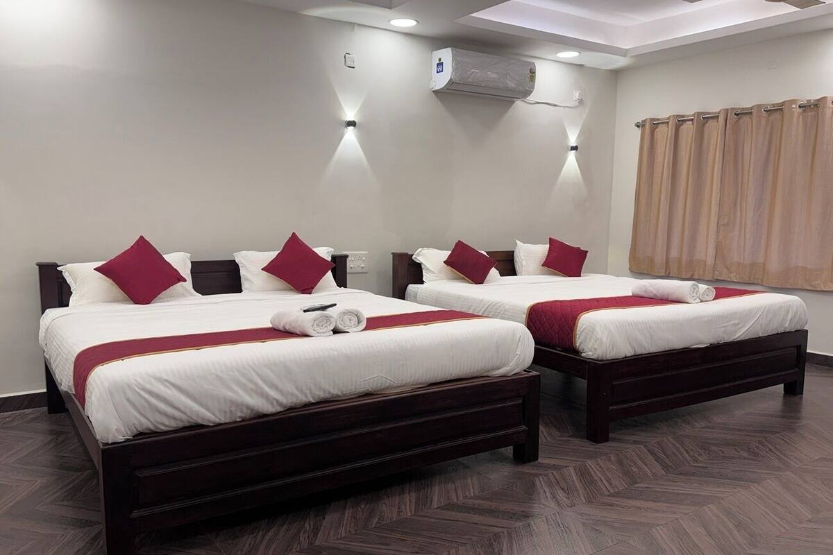 A hotel room with two beds, each with a wooden frame and white bedding, red pillows, and a red throw blanket. There is a wall-mounted air conditioning unit and a ceiling light fixture.