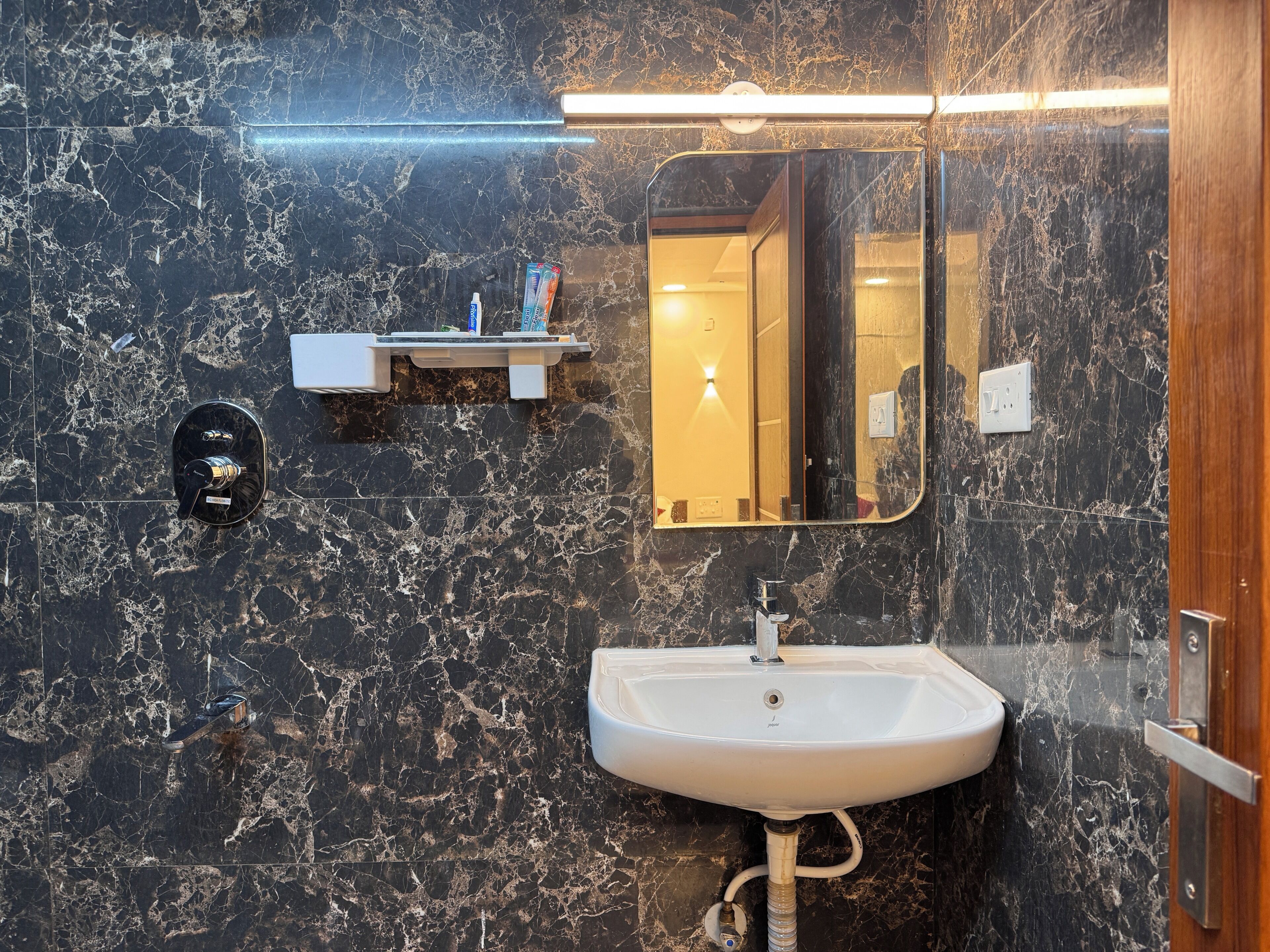A bathroom with a marble wall, a white sink, a mirror, and a shelf with toiletries.