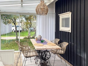 A covered patio with a wooden table and wicker chairs, a hanging light fixture, and a view of a backyard.