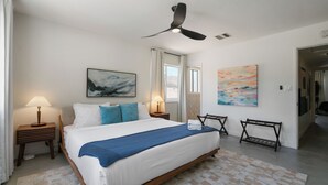 Une chambre d’hôtel comprenant un lit, deux tables de chevet, un ventilateur de plafond, une fenêtre avec des rideaux et des œuvres d’art accrochées aux murs.