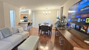 A modern living room with a sofa, ottoman, dining table, and chairs. A TV is mounted on the wall displaying various channels. - Downtown Chanhassen Townhome - 3 bedrooms (Chanhassen)
