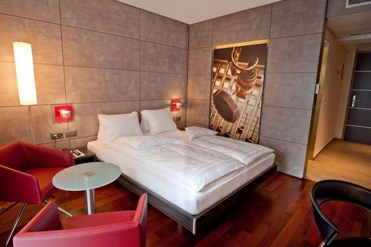 A hotel room with a bed, a small table, a red chair, and a wall-mounted artwork.