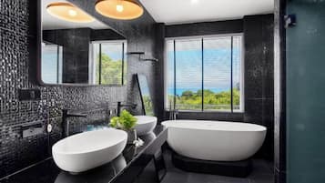 A modern bathroom with a large bathtub, two sinks, a large mirror, and a view of greenery through the window.