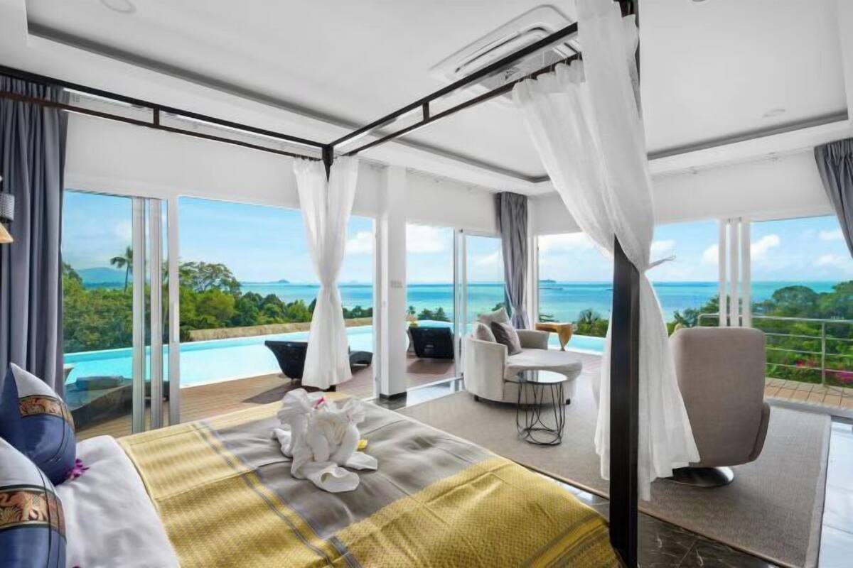 A bedroom with a large bed, a sitting area with a view of the ocean, and a balcony with a pool.