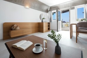 A modern room with a wooden headboard, a dining table with a book and a cup of coffee, and a view of the outdoors through large windows. - Santorini Princess Spa Hotel (Santorini)