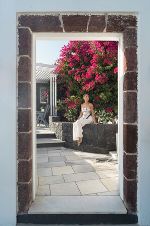A person sitting on a stone wall in a doorway framed by brick and white walls, with a view of a patio and a building in the background. - Santorini Princess Spa Hotel (Santorini)