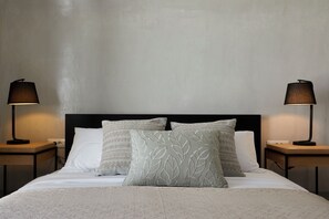 A neatly made bed with a white comforter and patterned pillows, flanked by two bedside tables with lamps. - Santorini Princess Spa Hotel (Santorini)