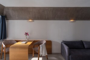 A modern room with a wooden dining table, two chairs, a dark sofa, and two wall-mounted lights. - Santorini Princess Spa Hotel (Santorini)