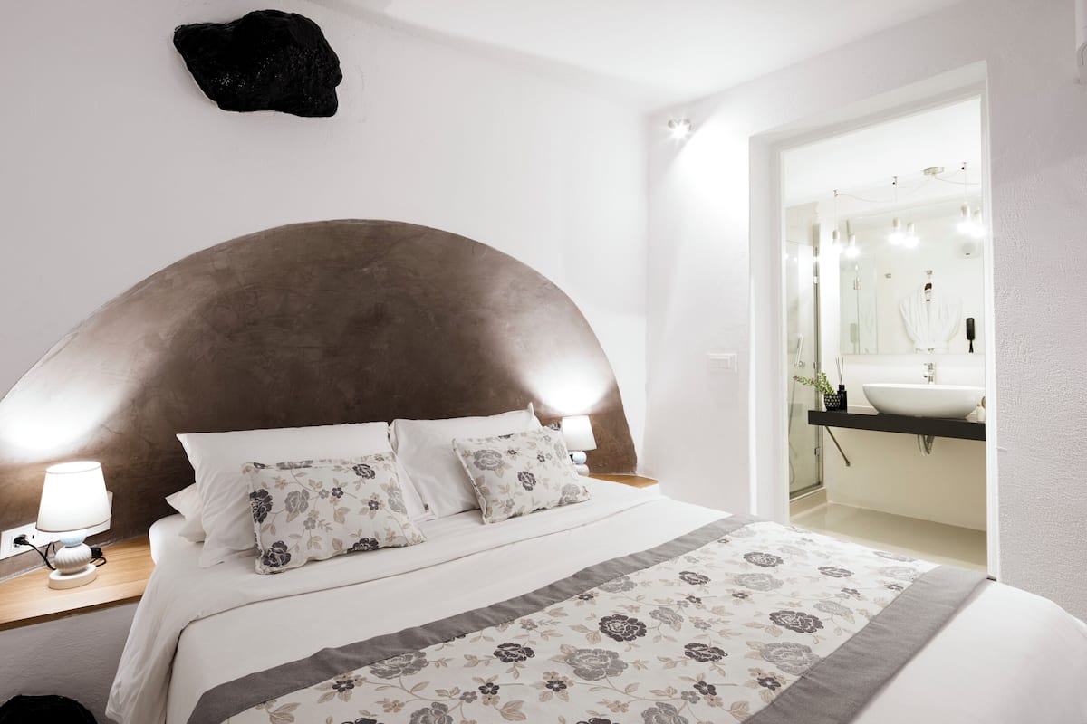 A bedroom with a large bed, a nightstand with a lamp, a mirror, and a bathroom visible through an open door.
