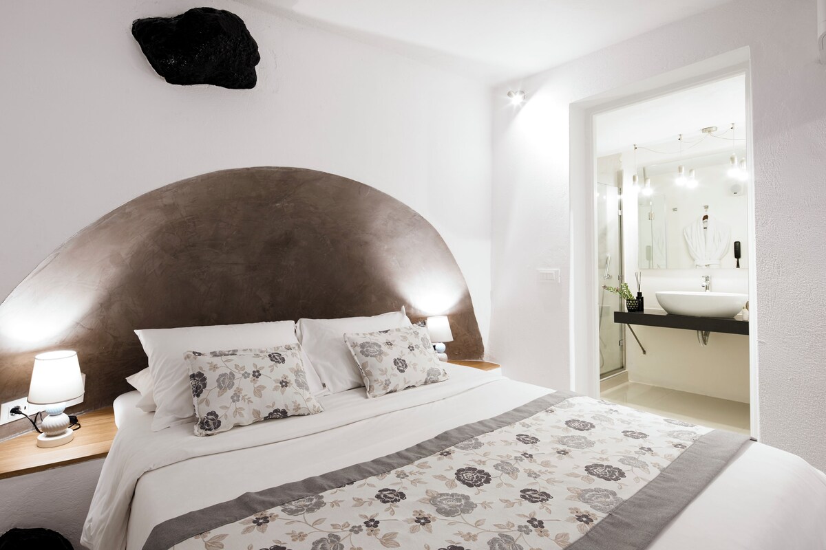 Un dormitorio con una cama grande, una mesita de noche con lámpara, un espejo y un baño visible a través de una puerta abierta.