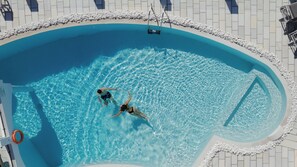 An aerial view of a swimming pool with two people swimming, surrounded by a pebble border and tiled deck.