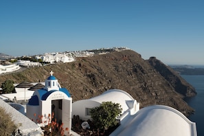 A white building with a blue dome and a cross, surrounded by a rocky landscape and a body of water.